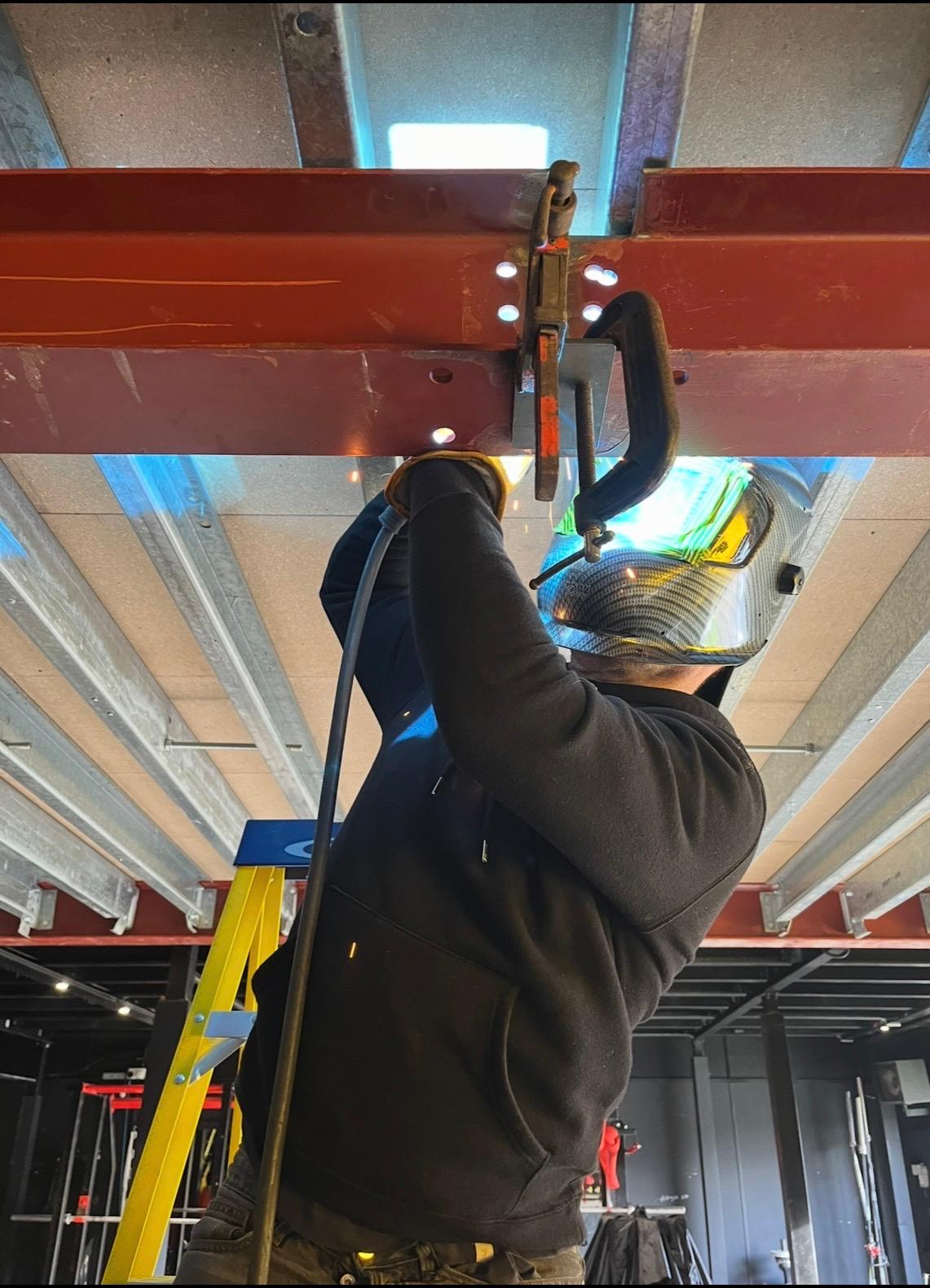 A welder joining a steel beam to a connection plate beneath a mezzanine deck